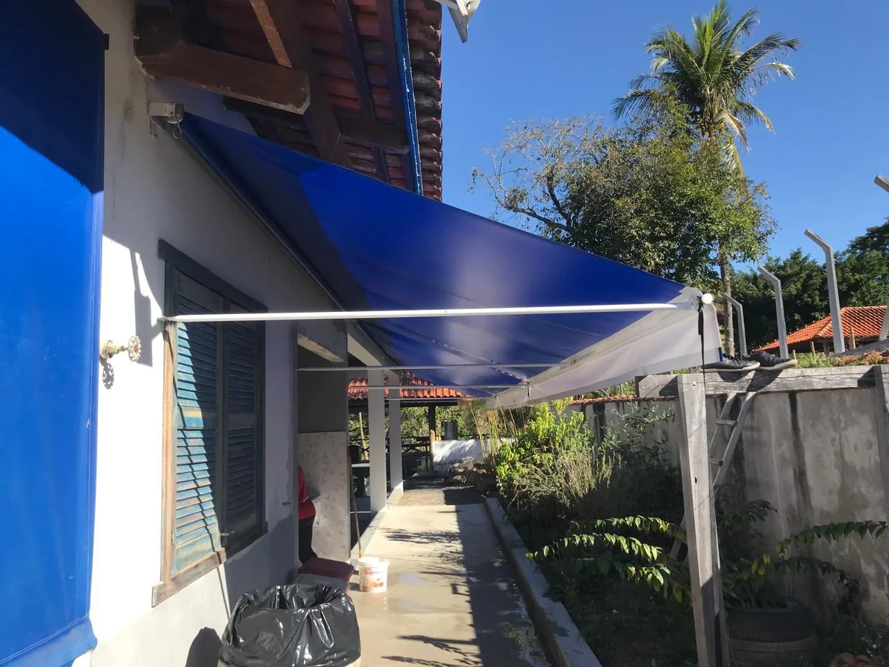 Toldo retrátil com braços azul aberto sobre a passagem lateral de uma casa